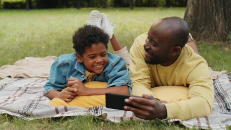 Afroamerikanischer-Vater-Und-Kleiner-Sohn-Liegen-Im-Park,-Telefonieren-Und-Chatten